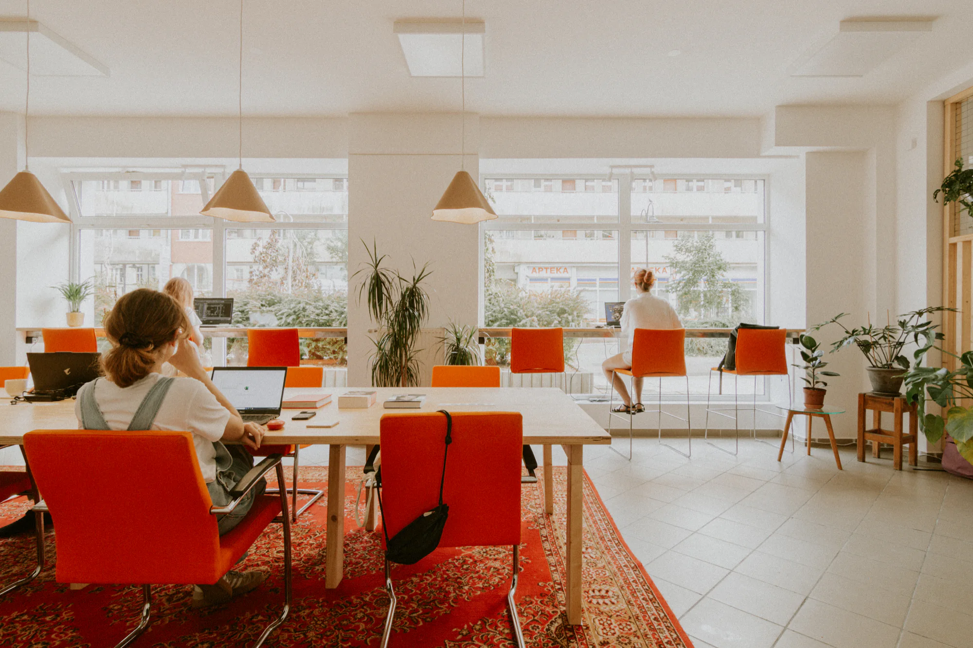 Wnętrze coworkingu ko_stacja: wspólny stół, pomarańczowe krzesła i stanowiska przy oknach z widokiem na ulicę Dworcową.